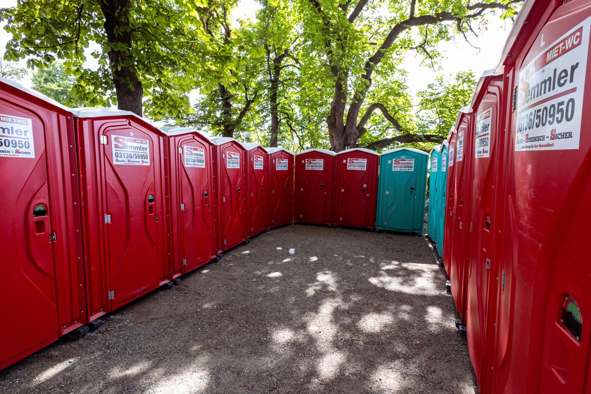 Red portable toilets lining a tree-shaded gravel path