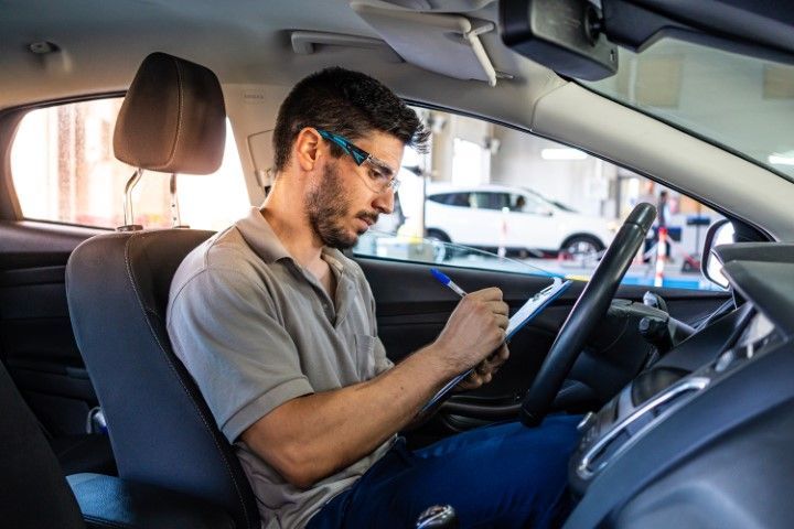 A person wearing glasses and a gray shirt sits in a car, writing on a clipboard while looking down.