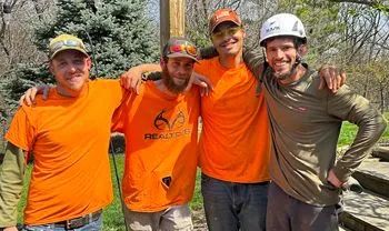Four men in work clothes, arms around each other, smiling in an outdoor setting. Most wear orange shirts; one wears a white helmet.