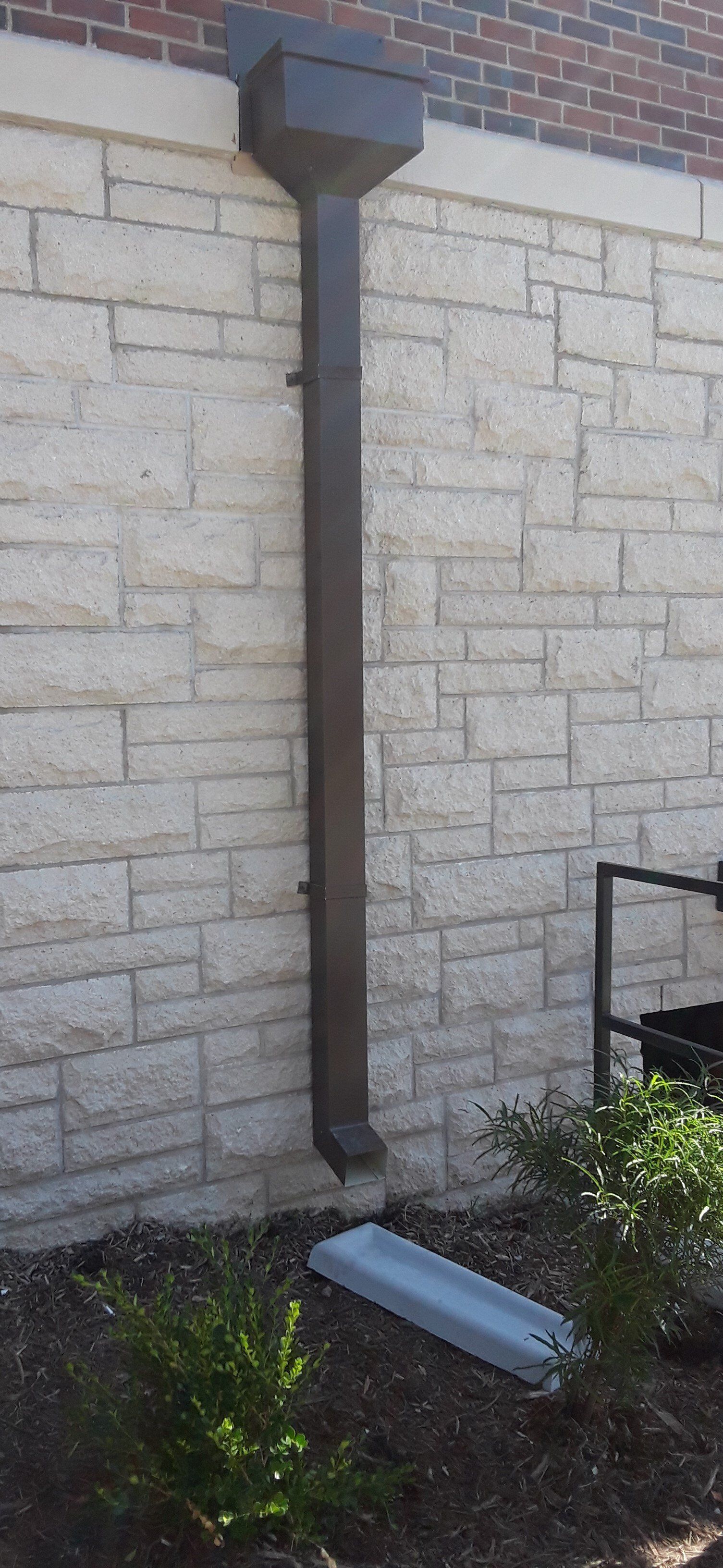Wall-mounted downspout pipe on a brick-and-stone building wall, with a fallen gutter section below.