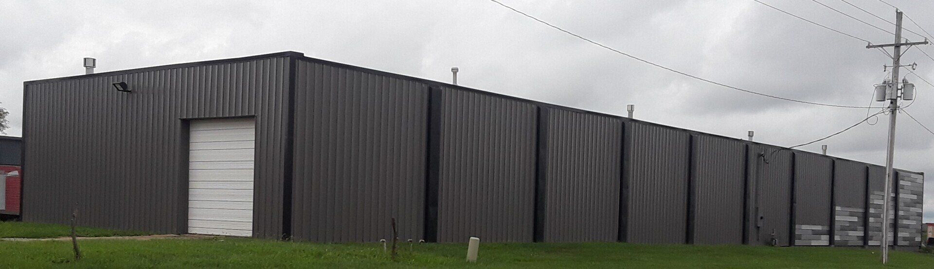 Long dark industrial building with a white garage door beside a grassy roadside under cloudy skies