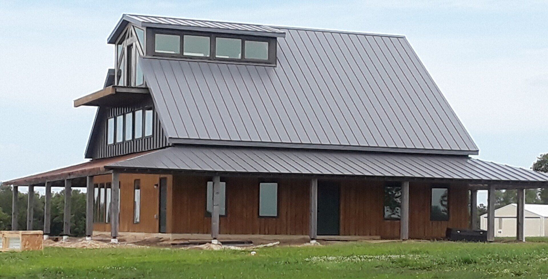 Modern brick house with a steep gray metal roof and covered wraparound porch in a grassy field