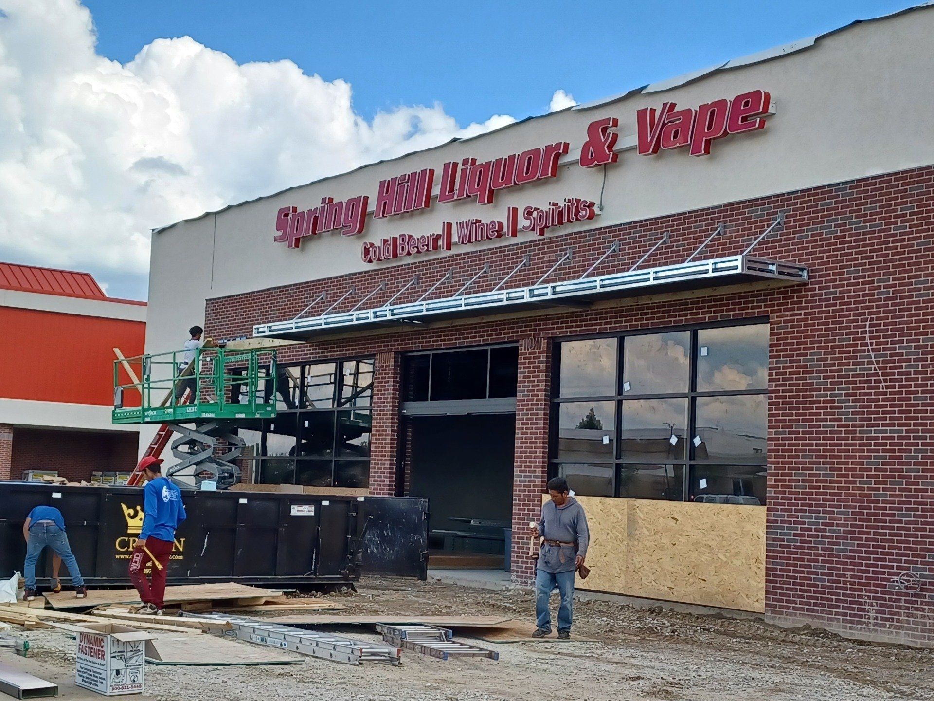Workers at a brick storefront under construction with red signage and scaffolding