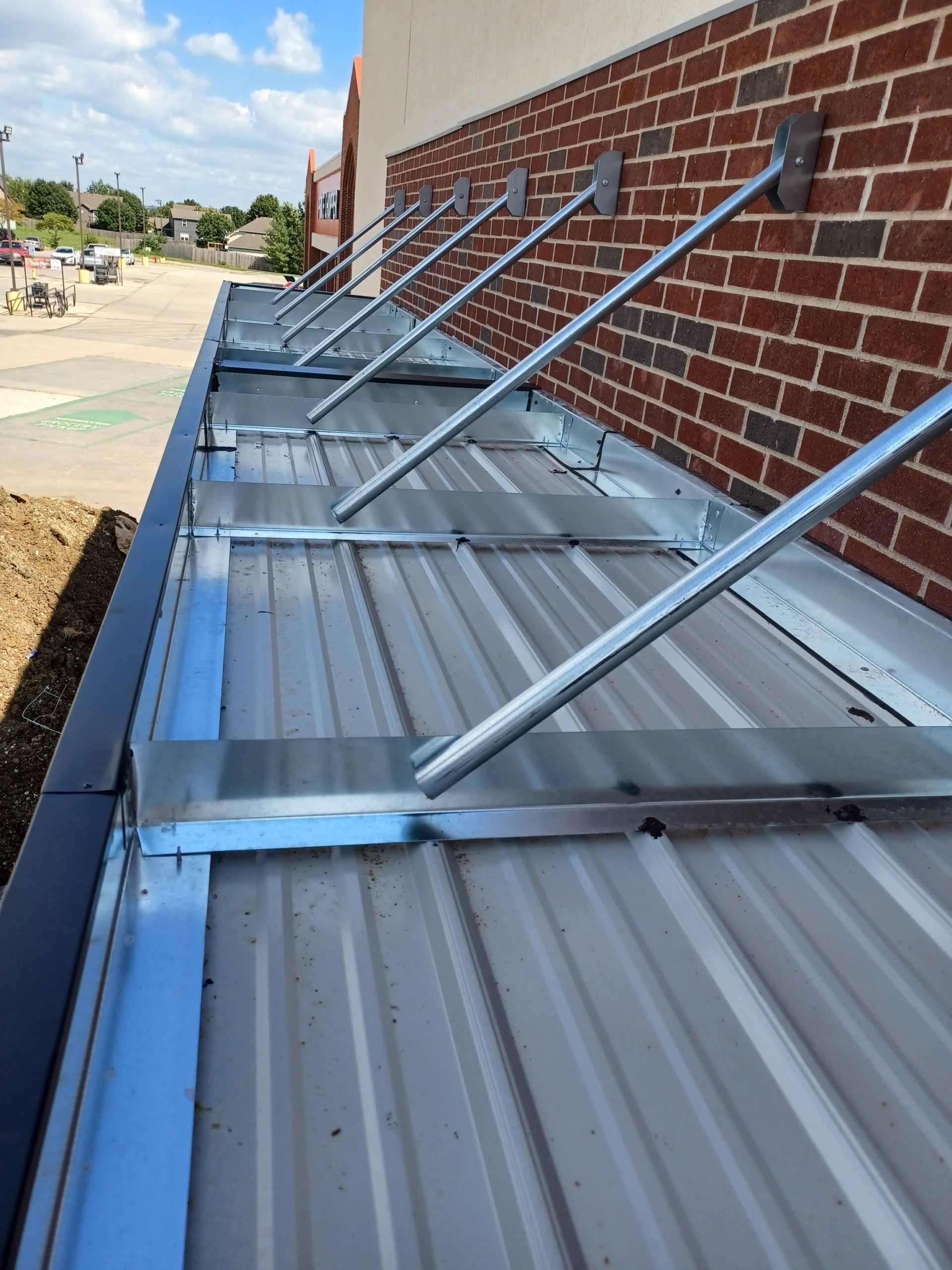 Blue metal roof panels and angled supports attached to a brick wall beside a sidewalk.