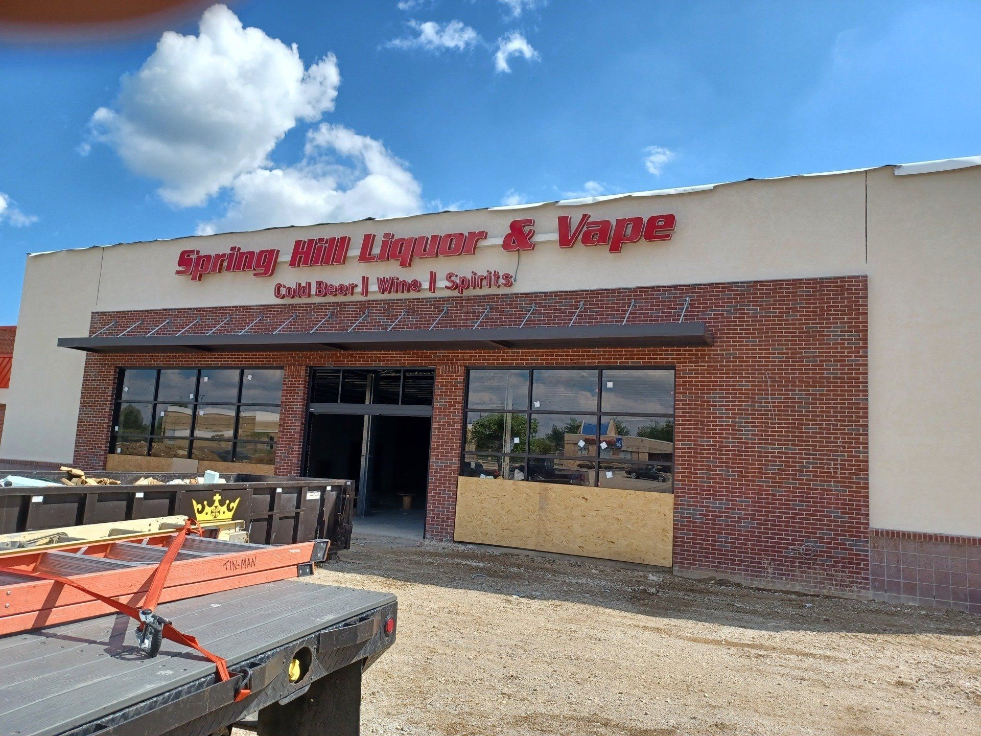 Storefront of a liquor and tobacco shop with red signage, glass doors, and a gravel lot under a blue sky.