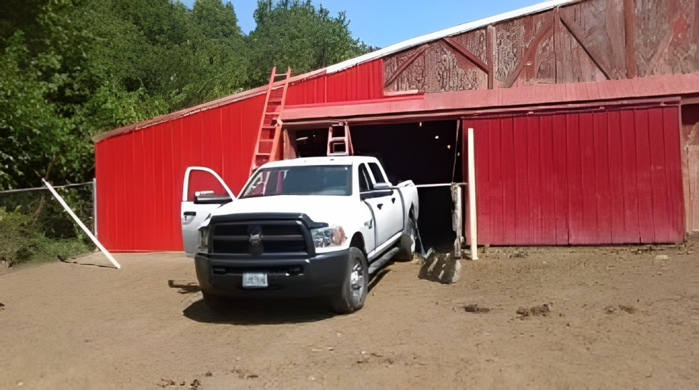 White pickup truck parked outside a red barn with open doors on a dirt lot.