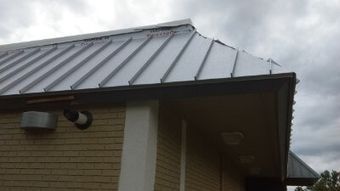 A gray metal roof on a brick house under a cloudy sky, with a vent and eave visible.