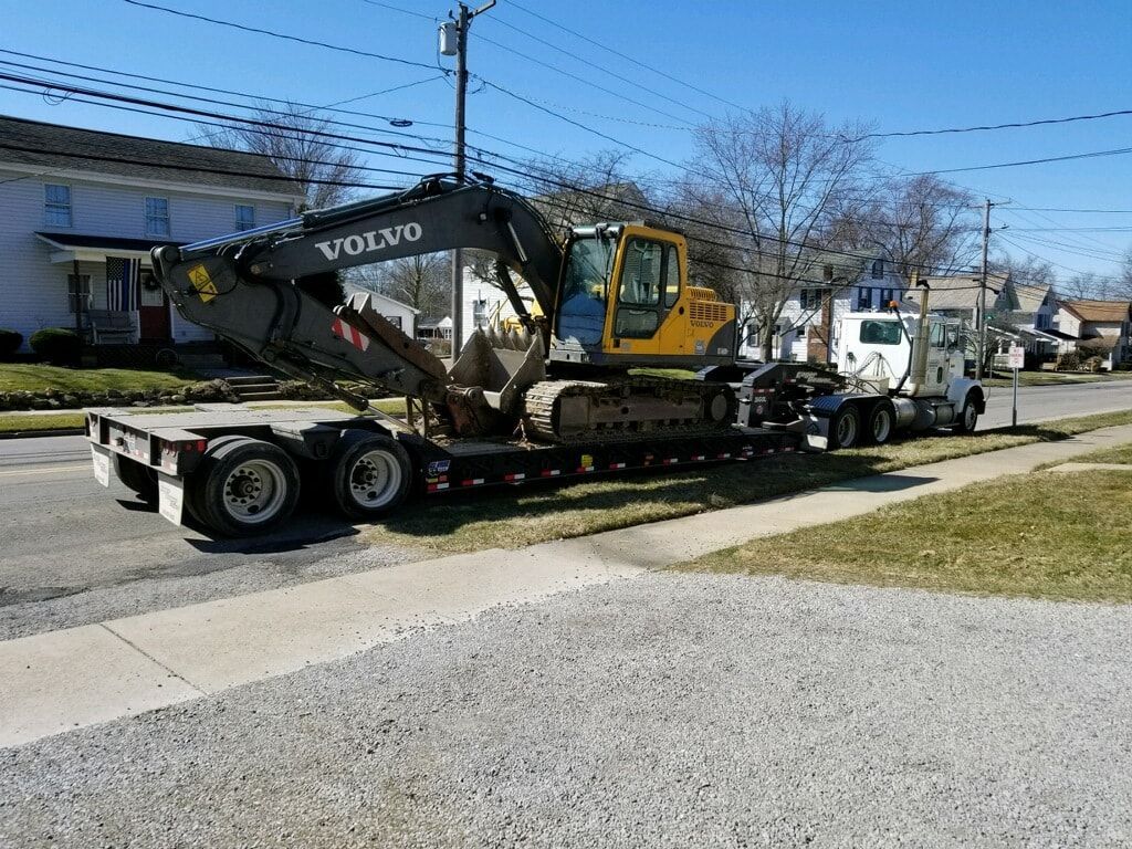 Volvo excavator on a flatbed trailer being transported by a white semi-truck on a residential street.