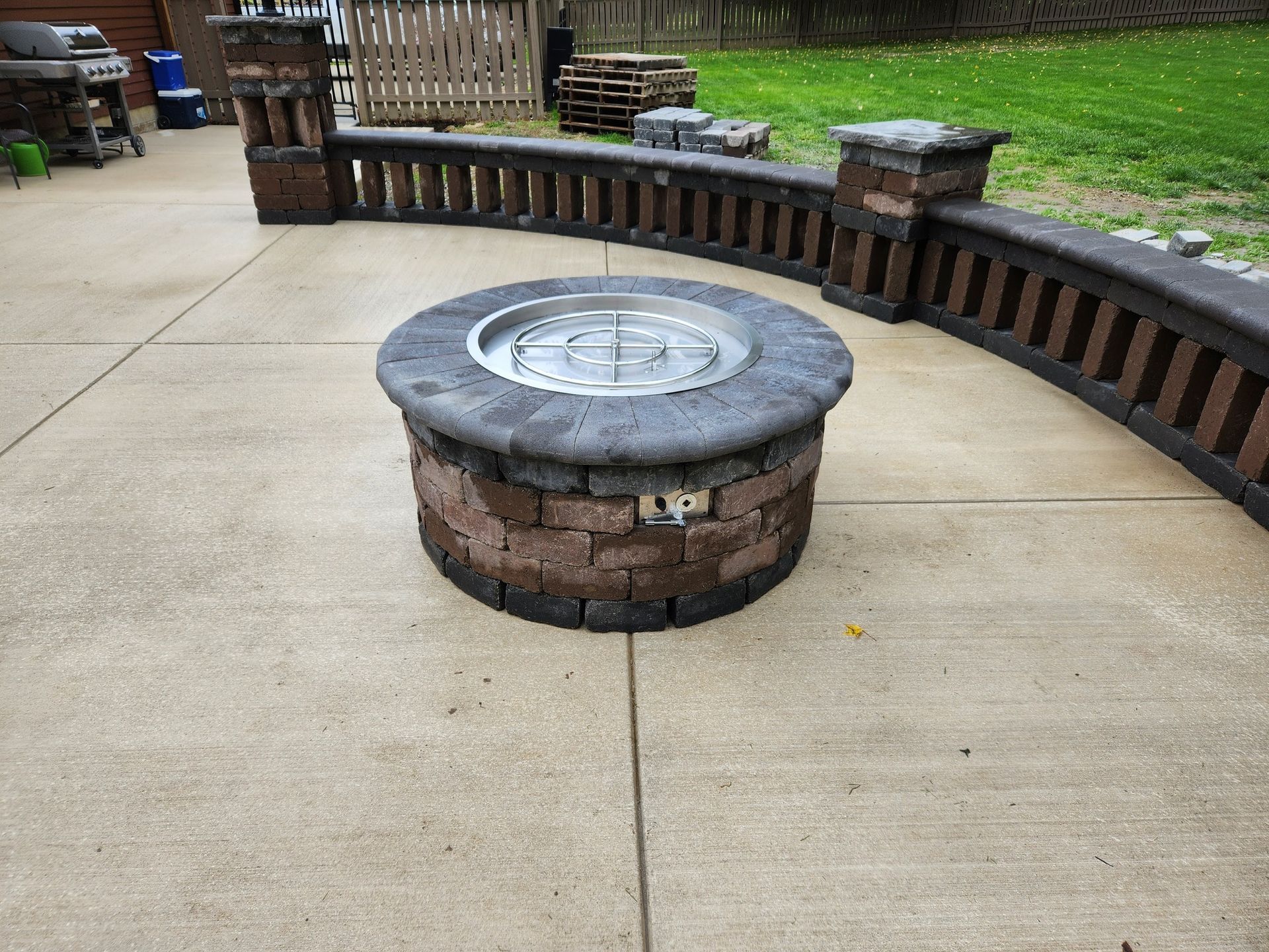 Circular brick fire pit on a concrete patio, with a surrounding brick wall and green grass in the background.