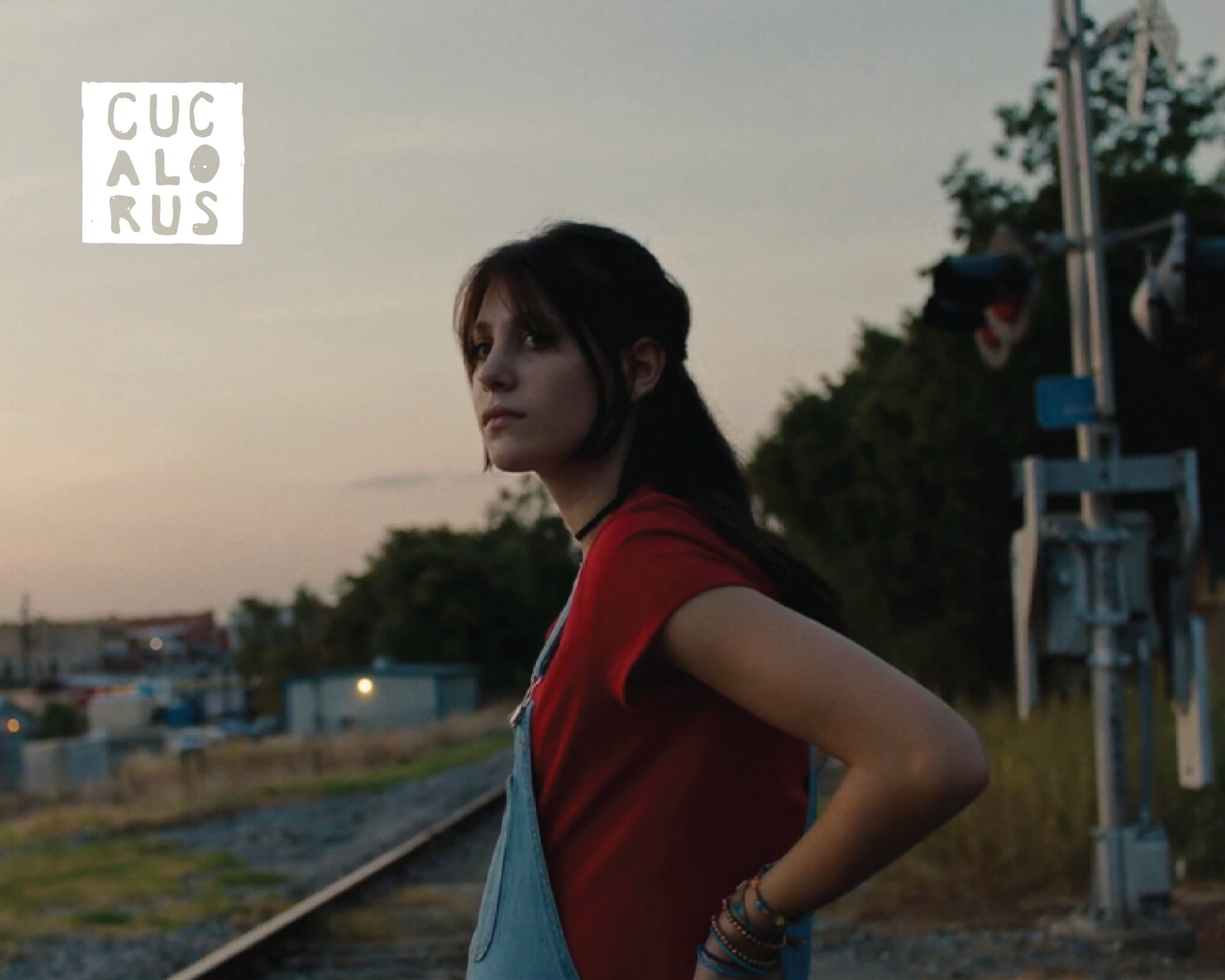 Woman in a red top standing by railroad tracks at sunset, looking into the distance.