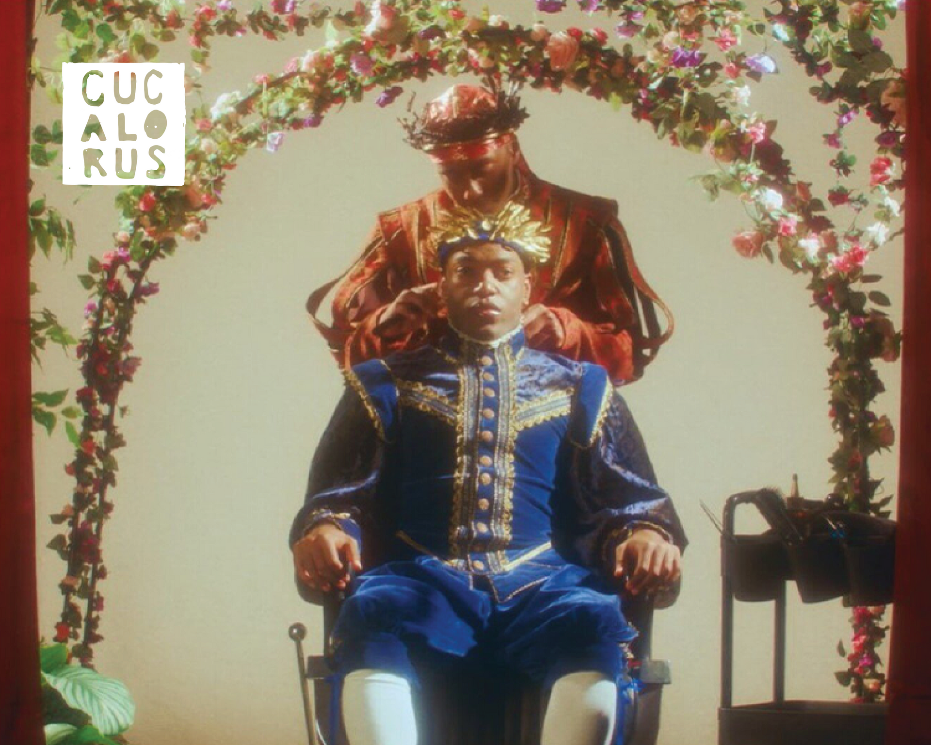 Two people seated beneath a floral arch, one in a blue outfit and one standing behind, in warm lighting