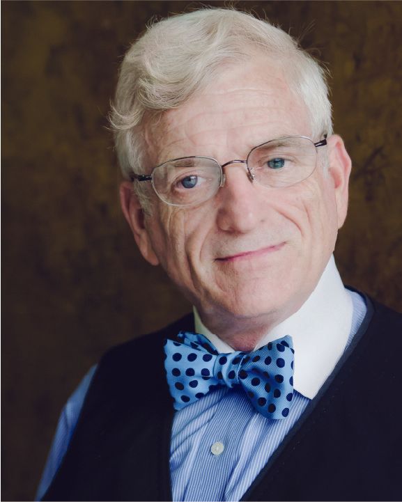 Portrait of a smiling person in glasses, blue shirt, and polka-dot bow tie against a dark background