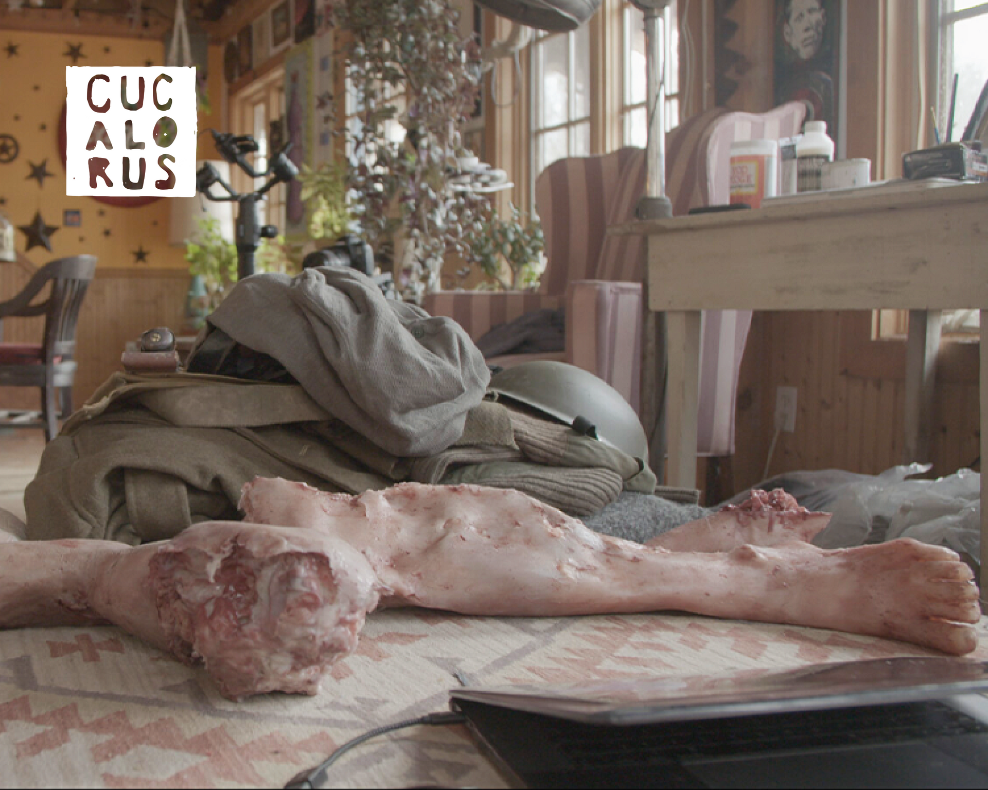Life-sized body sculpture on a table in a cluttered workshop with draped fabric and windows.