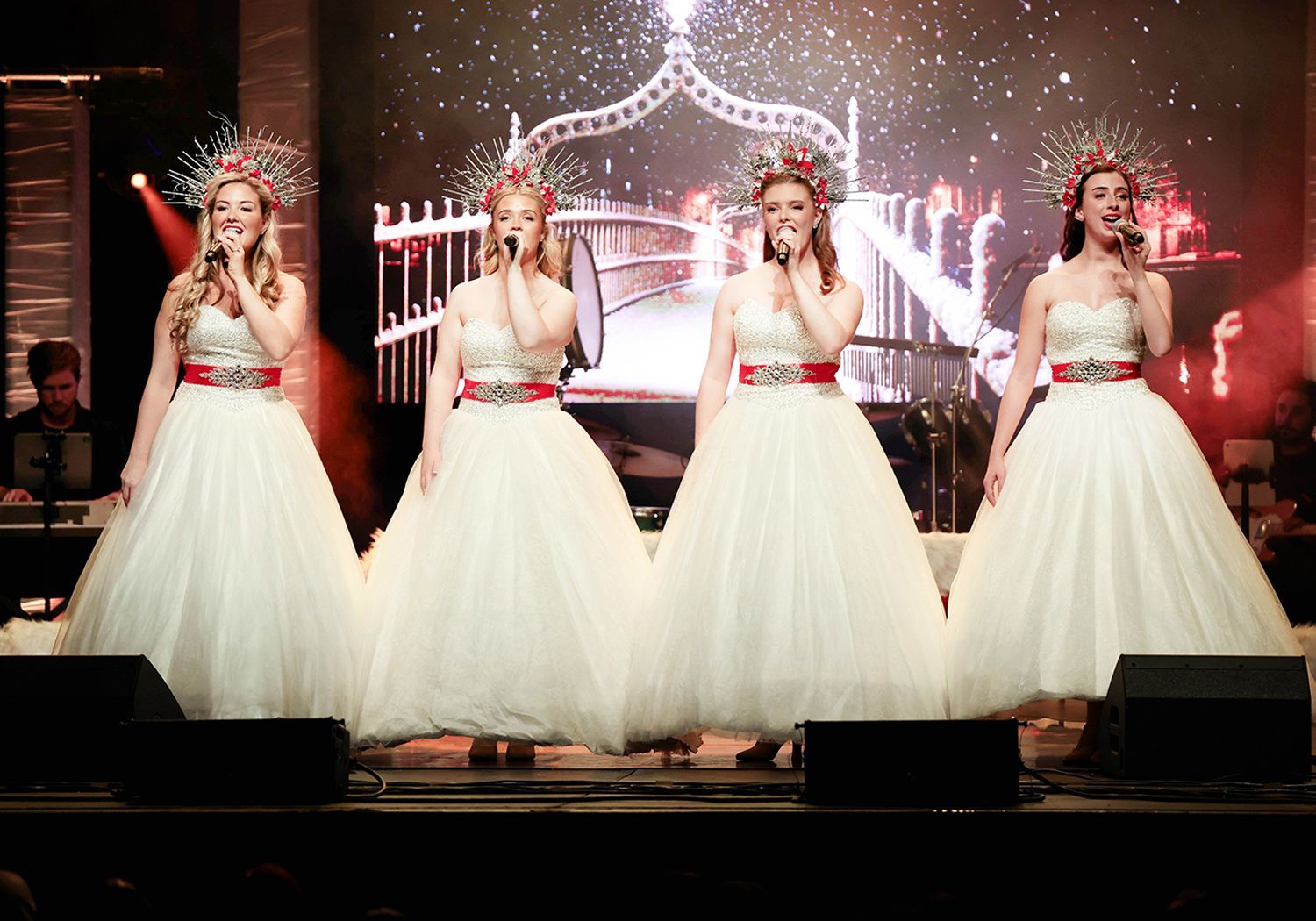 Four women in white gowns and crowns perform on a stage with pink lights and a chandelier backdrop