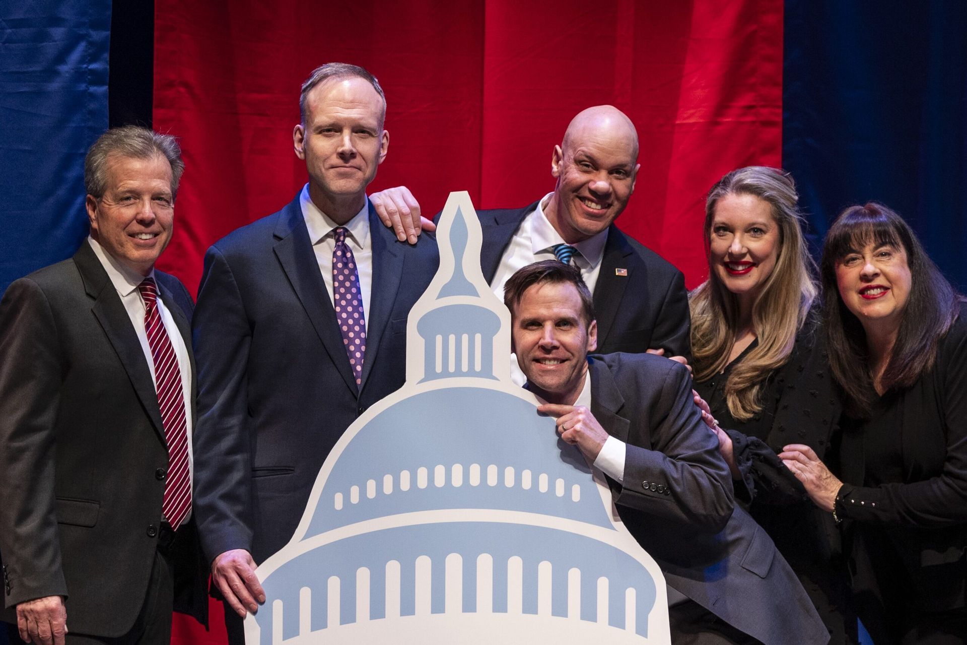Five people posing in front of a U.S. Capitol cutout, smiling on a stage with red curtains.