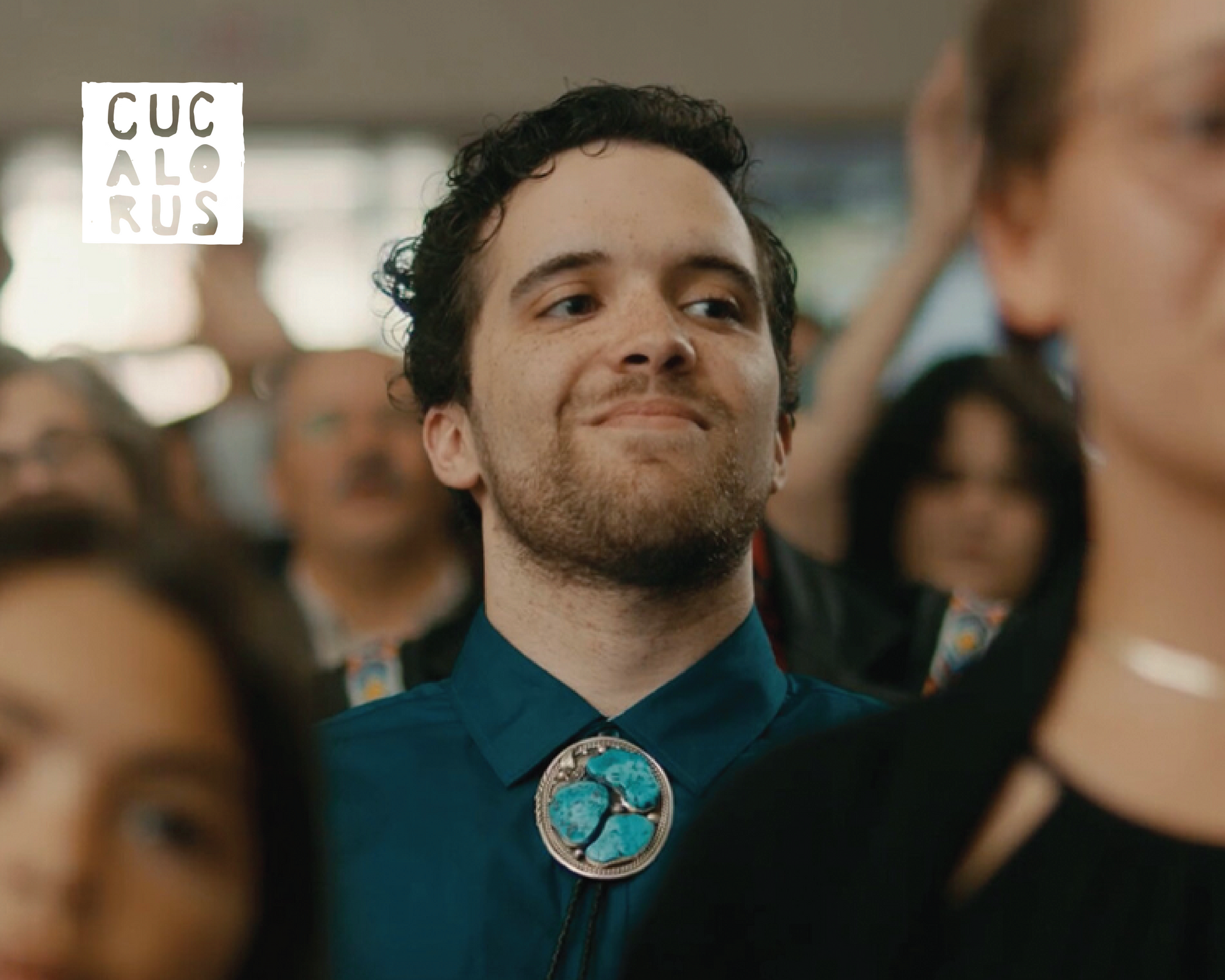 Smiling person in a teal shirt with a round turquoise pin, standing in a crowd at an event