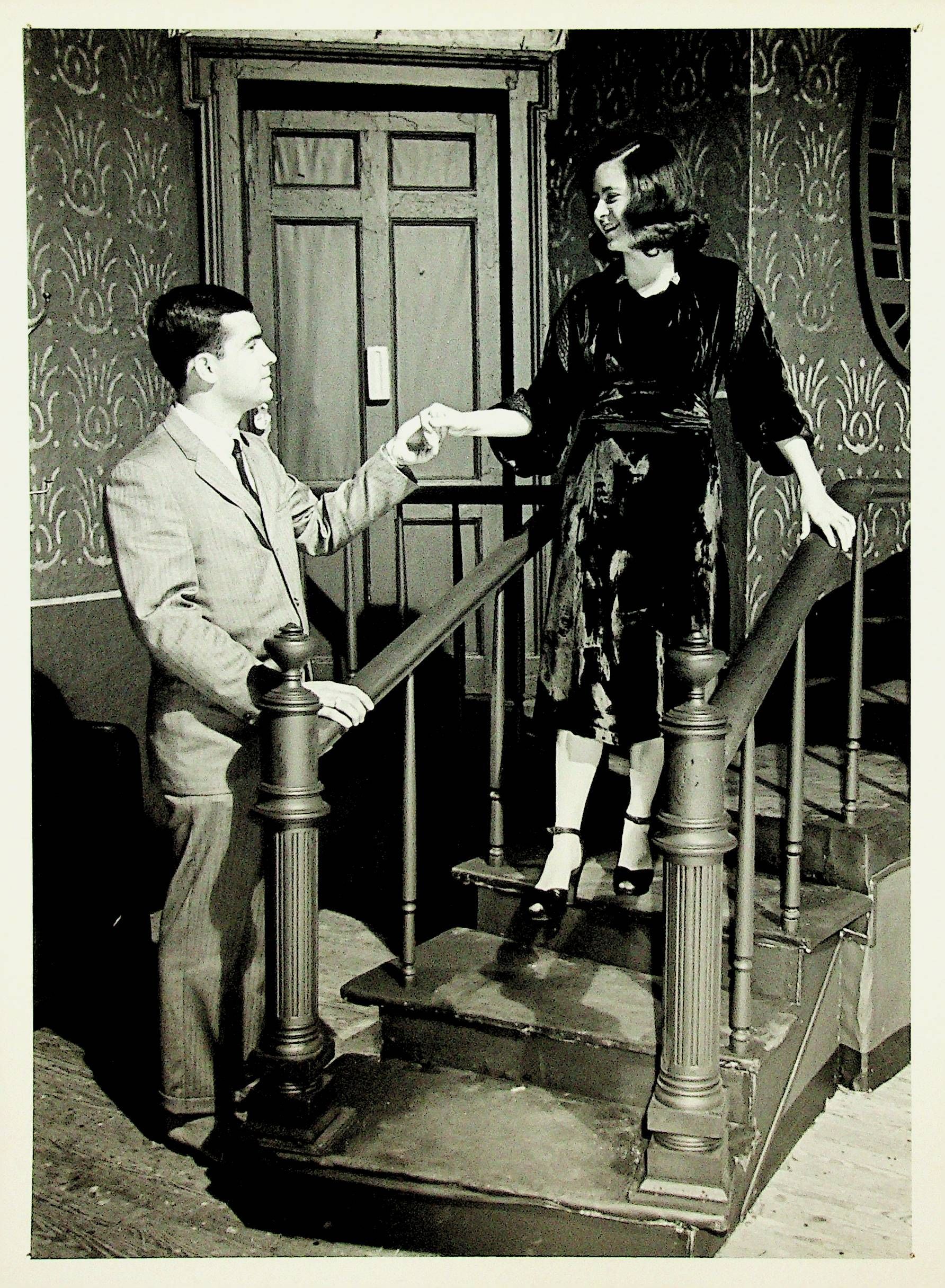 Man on a staircase offers his hand to a woman descending, in a narrow hallway.