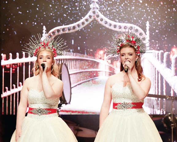 Four performers in white gowns sing onstage under pink lights, wearing ornate headpieces.