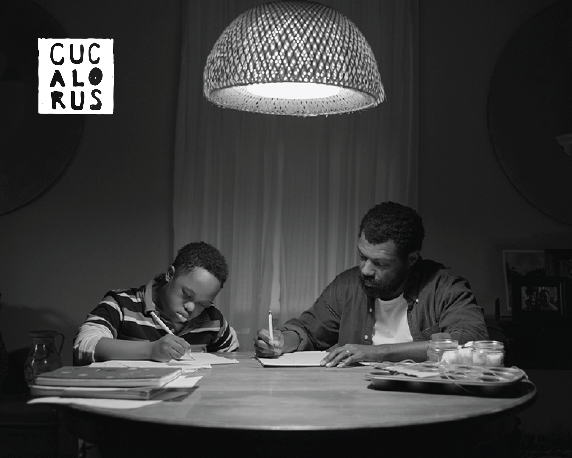 Two people writing at a table under a hanging lamp in a dim room