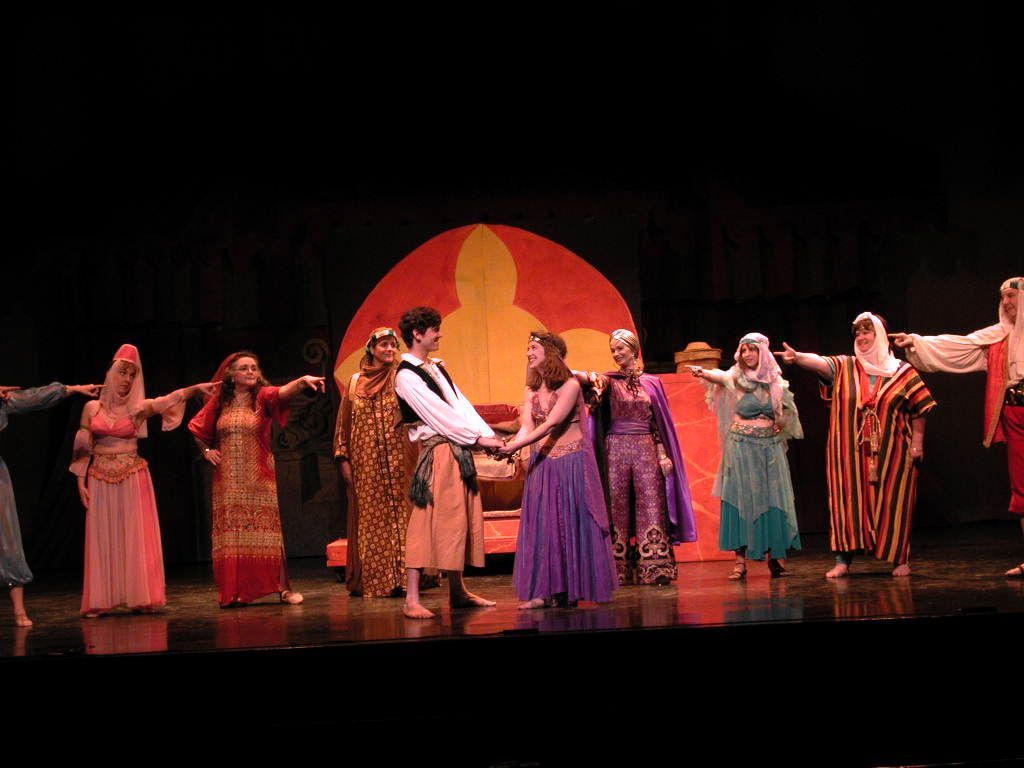 Dancers in colorful costumes hold hands on a stage before a large orange sun backdrop.