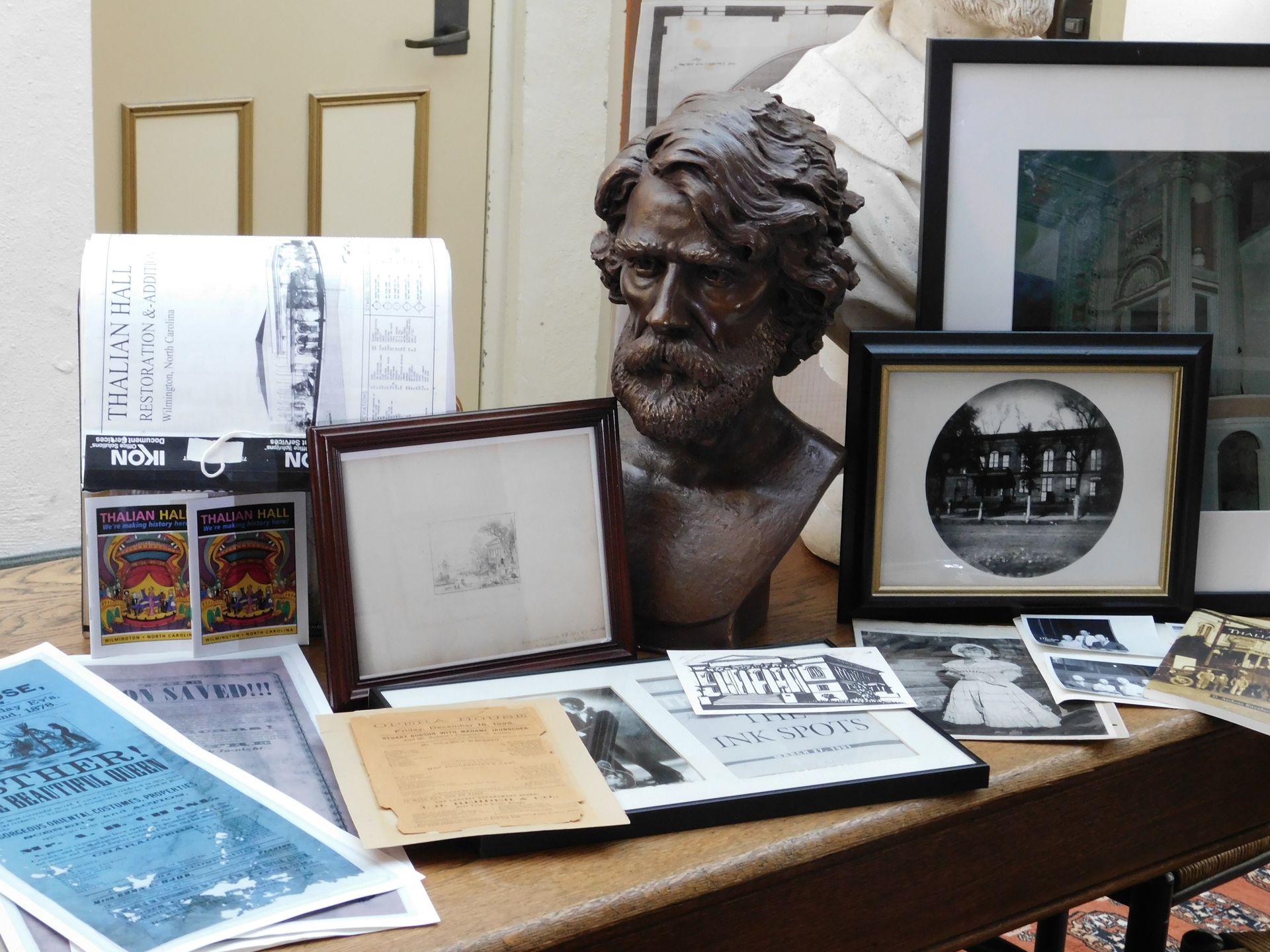 Display of framed artwork, books, and a bronze bust on a table in a gallery or museum setting