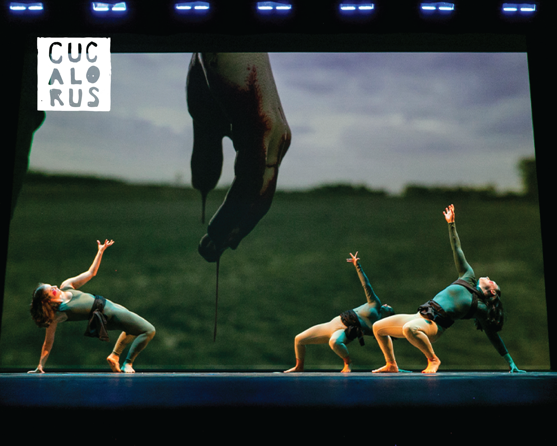 Three dancers in teal perform a bridge pose under a suspended figure on a stage.