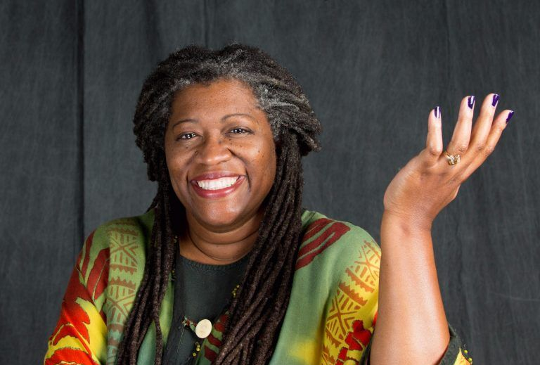 Smiling woman in a colorful patterned top, holding one hand up against a dark background