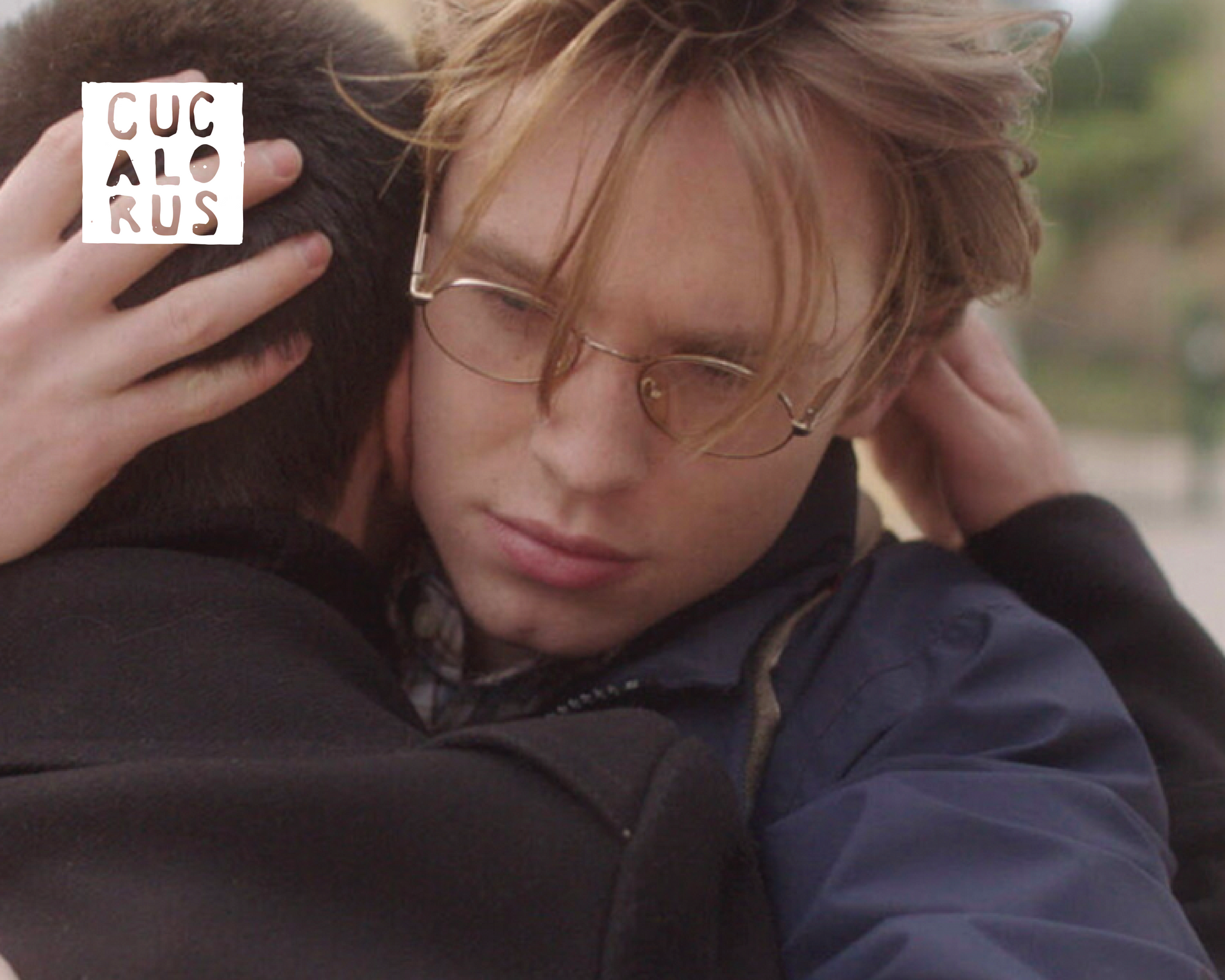 Two people embracing outdoors; one wears glasses and looks solemn
