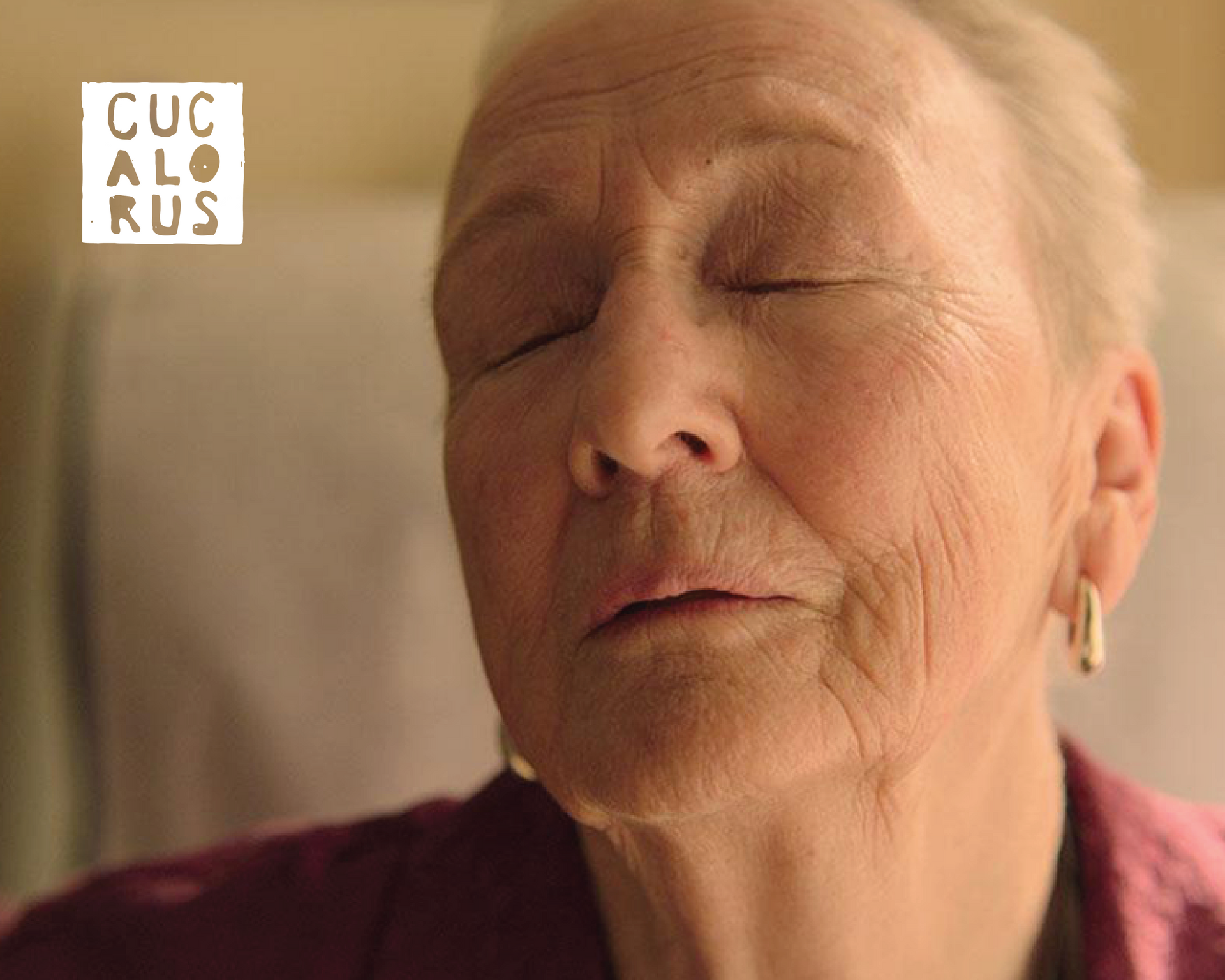 Close-up of an elderly person with eyes closed, wearing a maroon top, in warm indoor light.