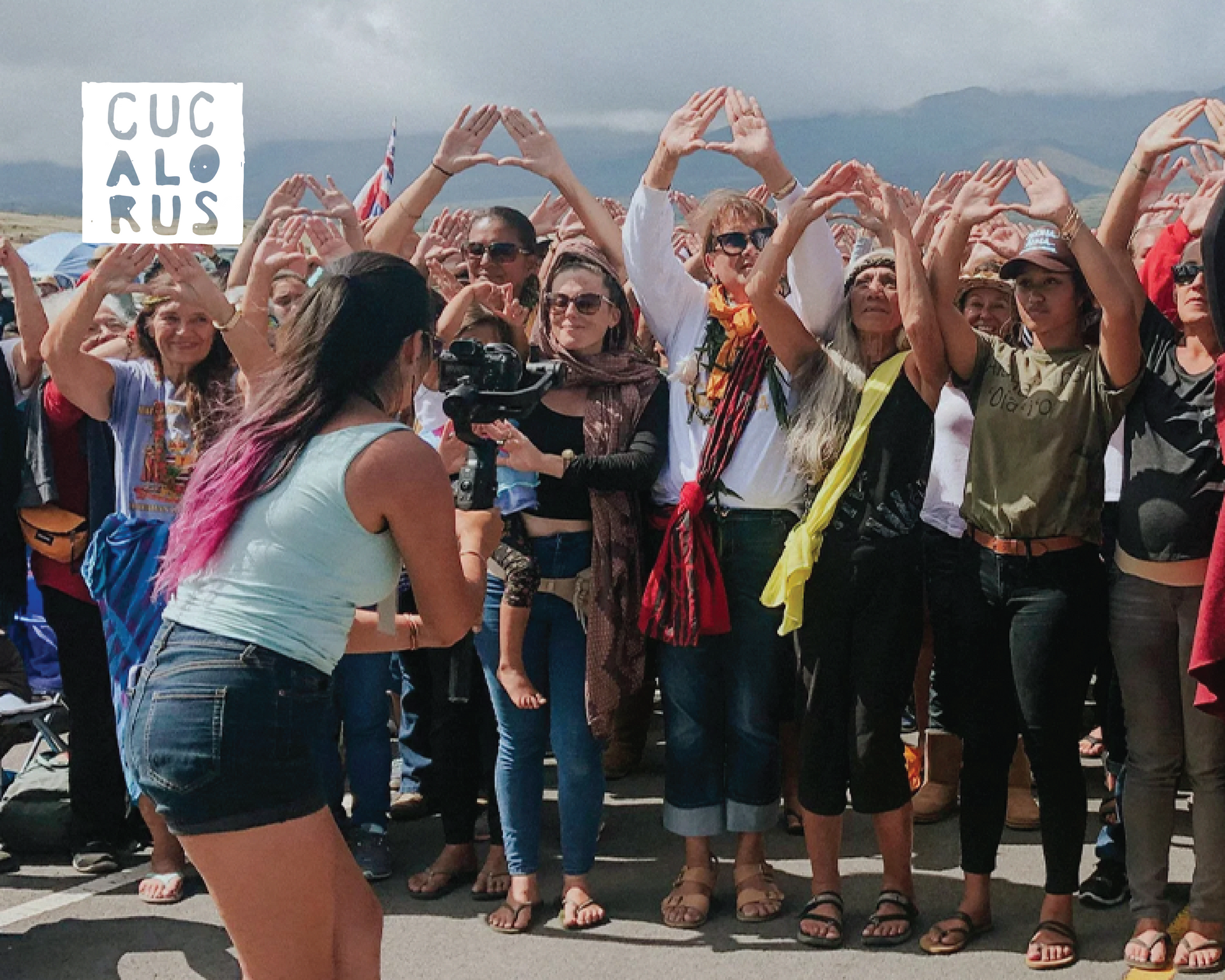 Group of people outdoors making heart shapes with their hands, smiling in a crowd near mountains.