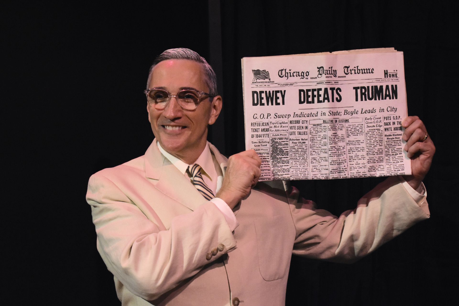 Man in a pink suit holding a newspaper reading “Dewey Defeats Truman,” smiling onstage