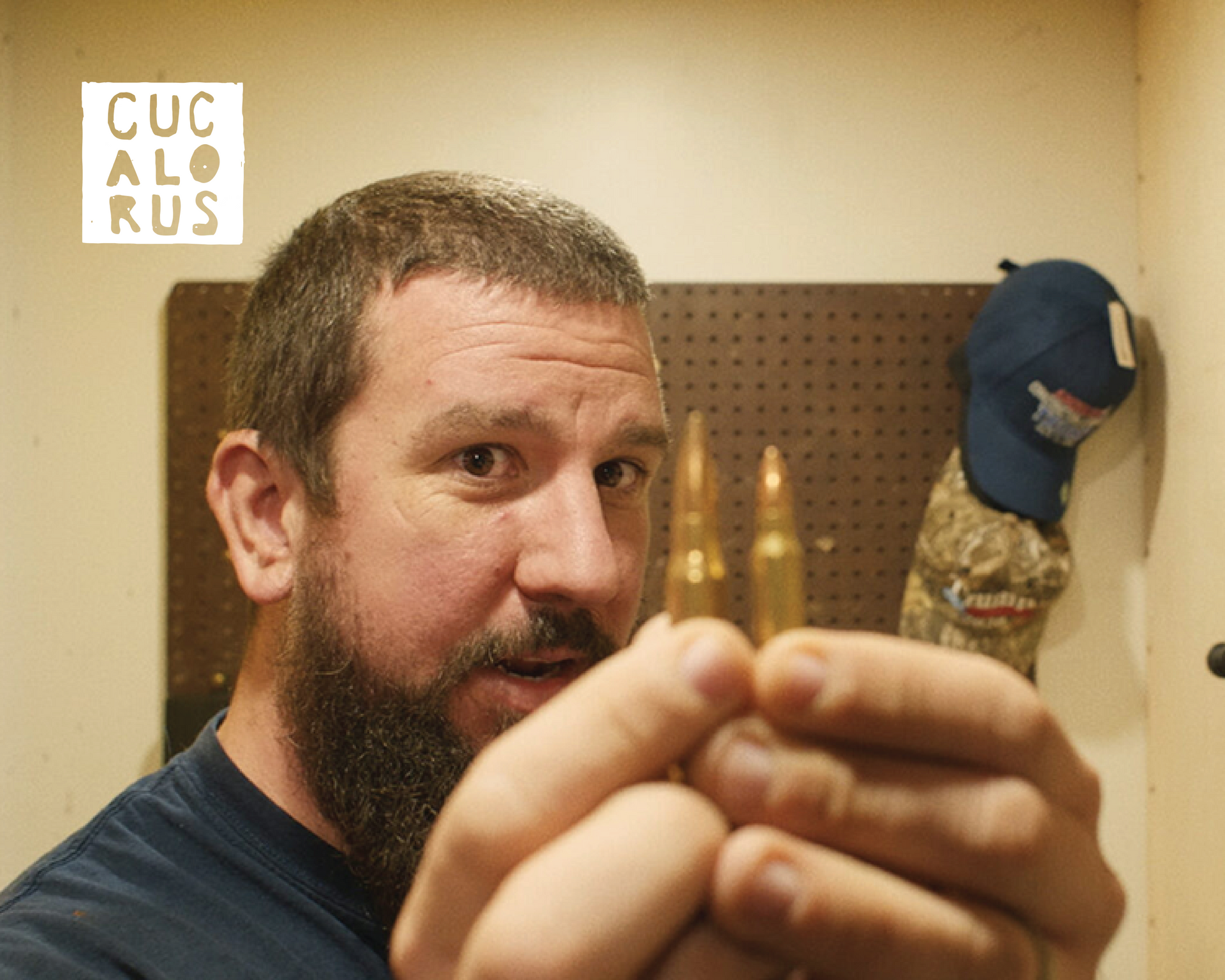 Man with a beard holding small wooden pieces toward the camera in a workshop; a blue cap hangs in the background.