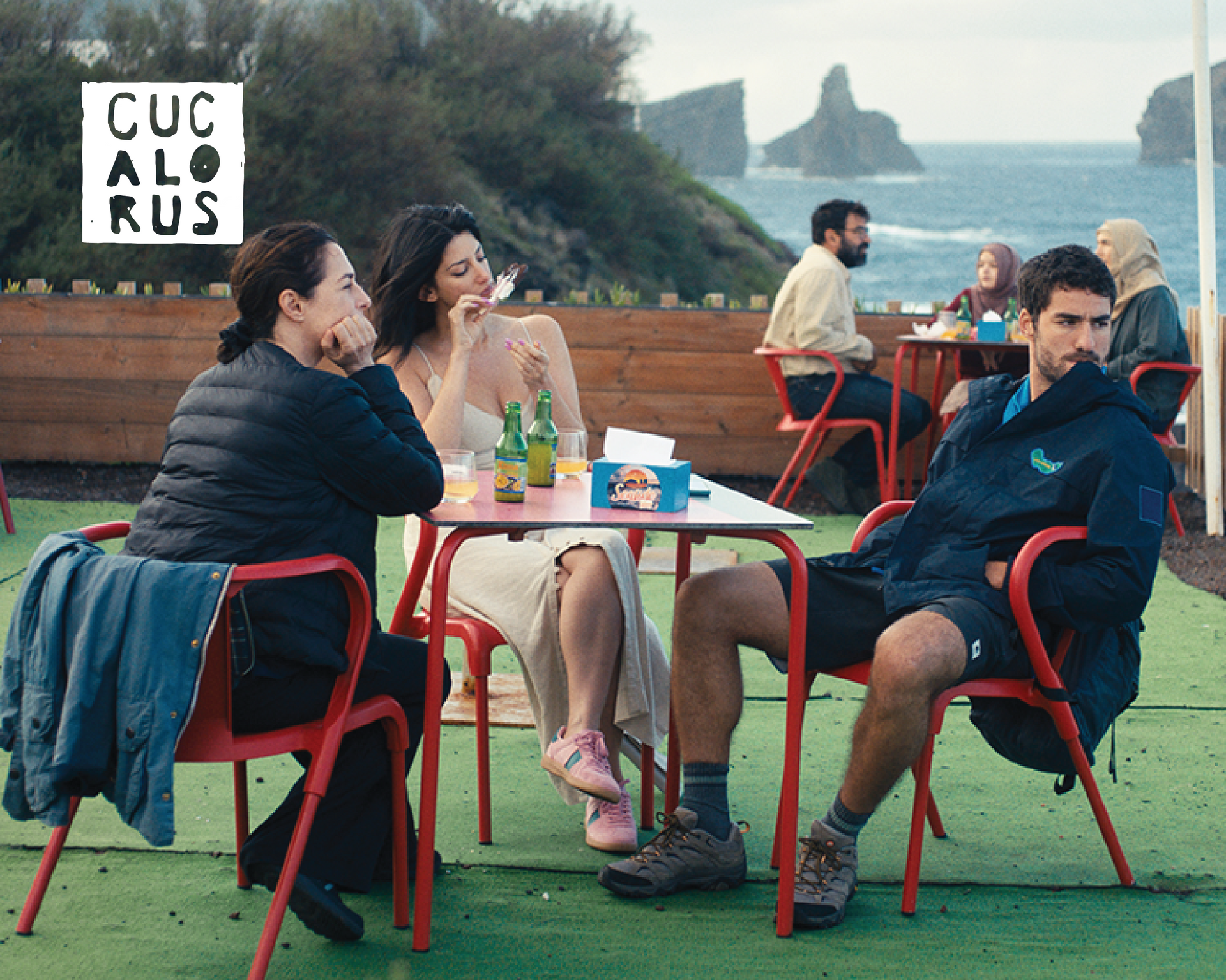 People sitting at red café tables on a grassy waterfront terrace, with a cloudy sea view.