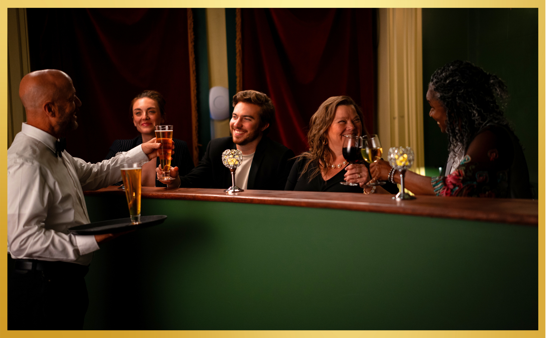 People toasting drinks at a bar, smiling around a green counter in warm low light