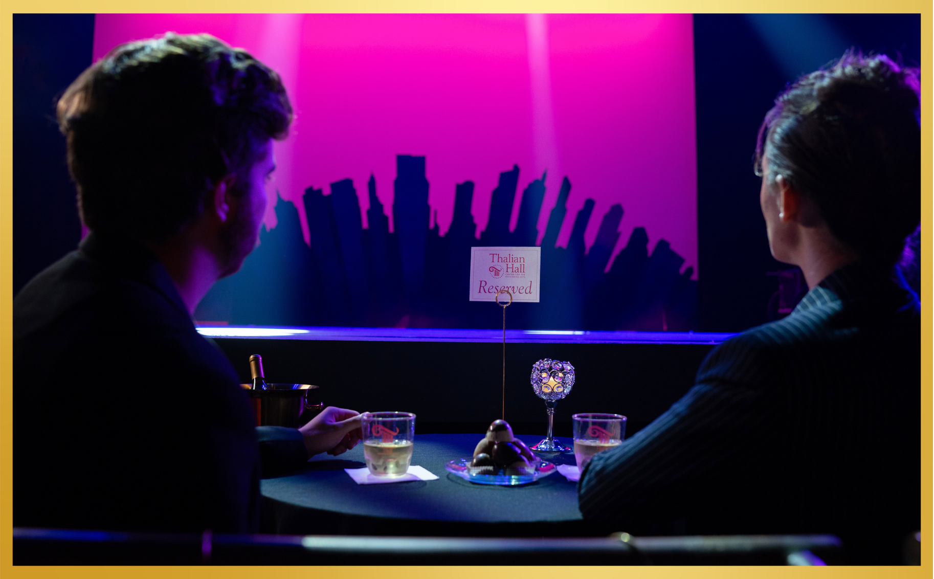 Two people seated at a dimly lit table, facing a neon-purple window with city silhouettes beyond.