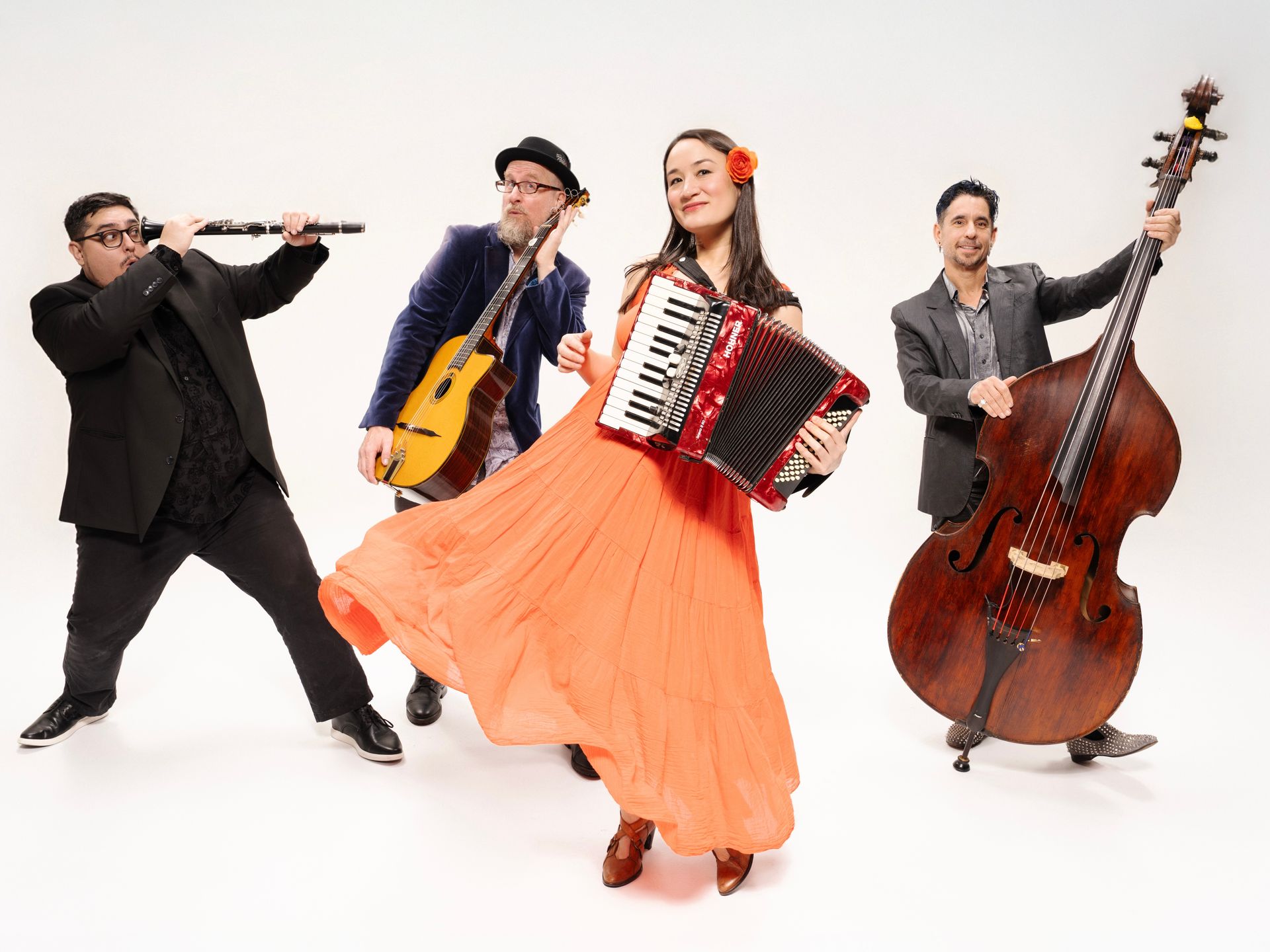 Four musicians perform on a white studio backdrop, with accordion, cello, flute, and double bass.