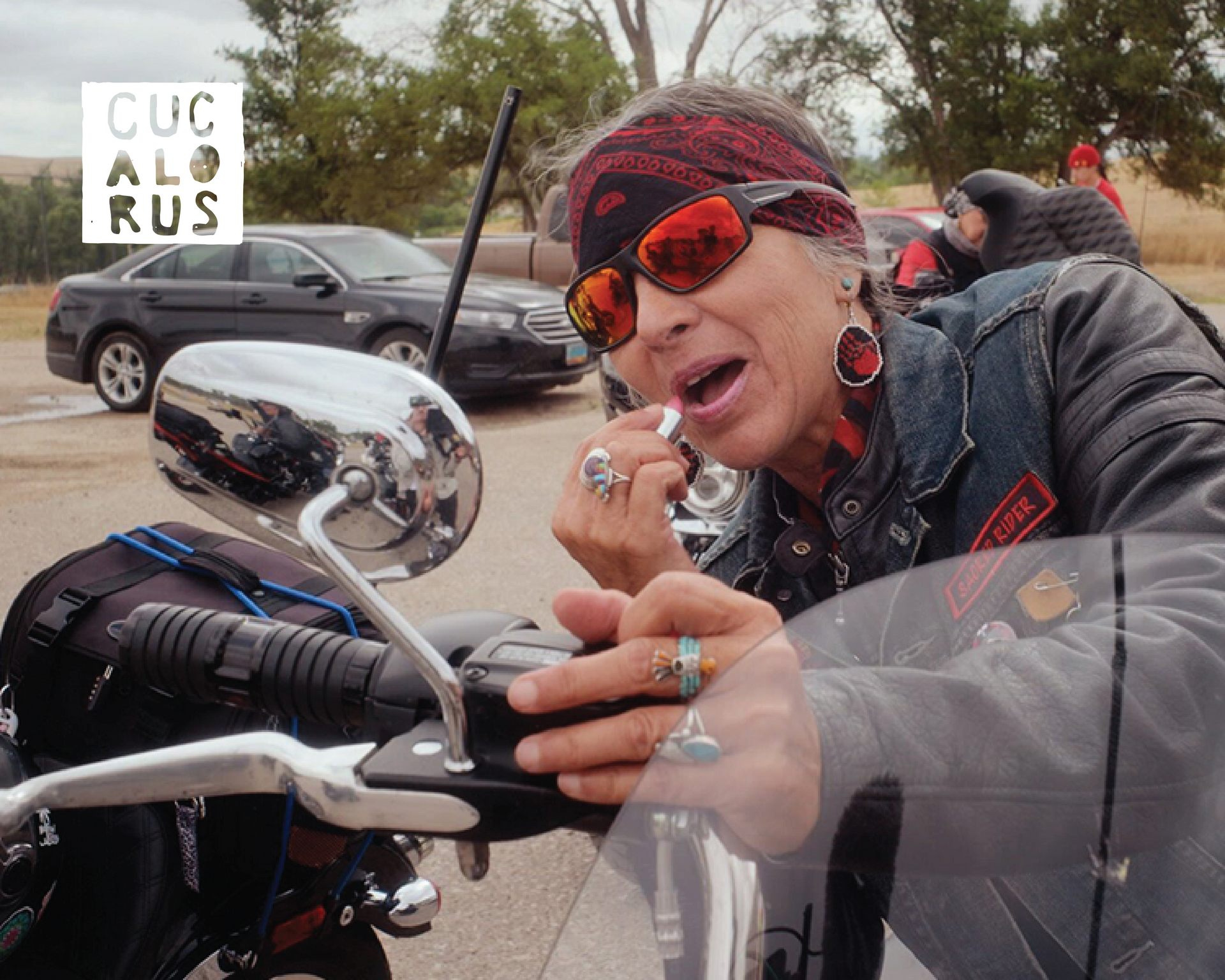Biker with red sunglasses and bandana beside a chrome motorcycle, with cars and a cloudy sky behind.
