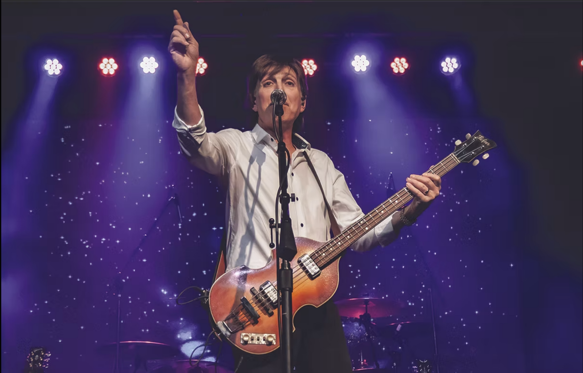Singer with electric guitar on stage, pointing upward under purple lights and a starry backdrop