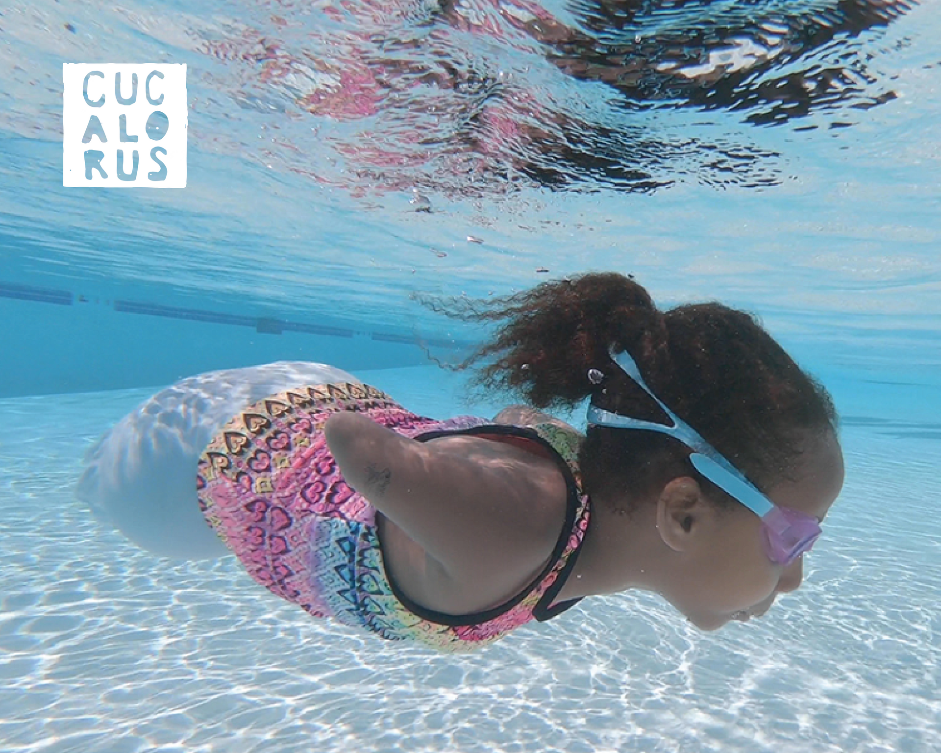 Girl swimming underwater in a pool, wearing pink goggles and a patterned swimsuit.