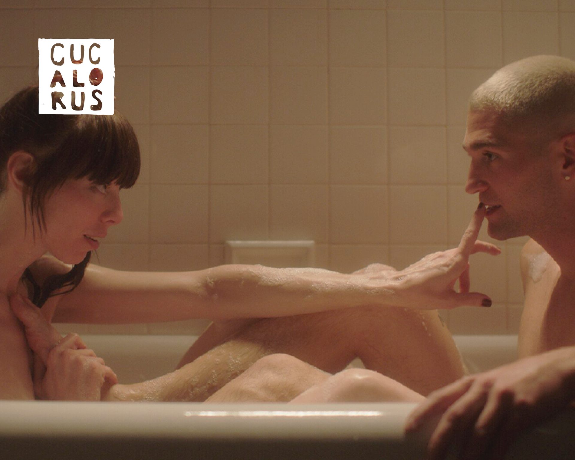 Two people relax in a bathtub, one reaching toward the other's face in a tiled bathroom.