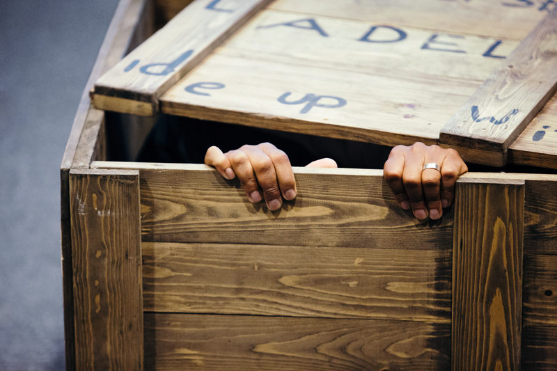 Hands gripping the edge of a wooden crate, with a partially visible person hiding inside