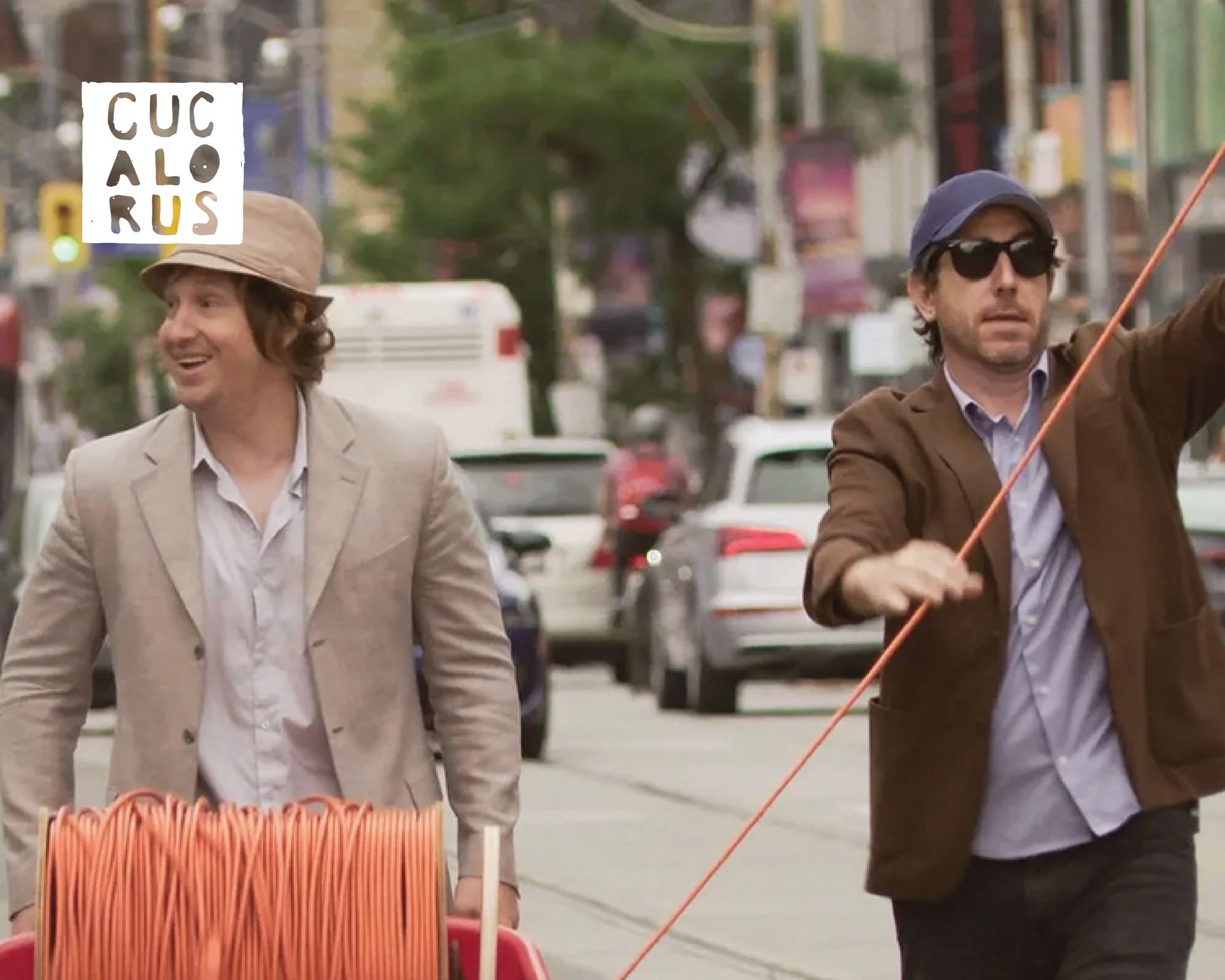 Two men walk down a city street, one pulling an orange cart and the other holding a long orange cable.