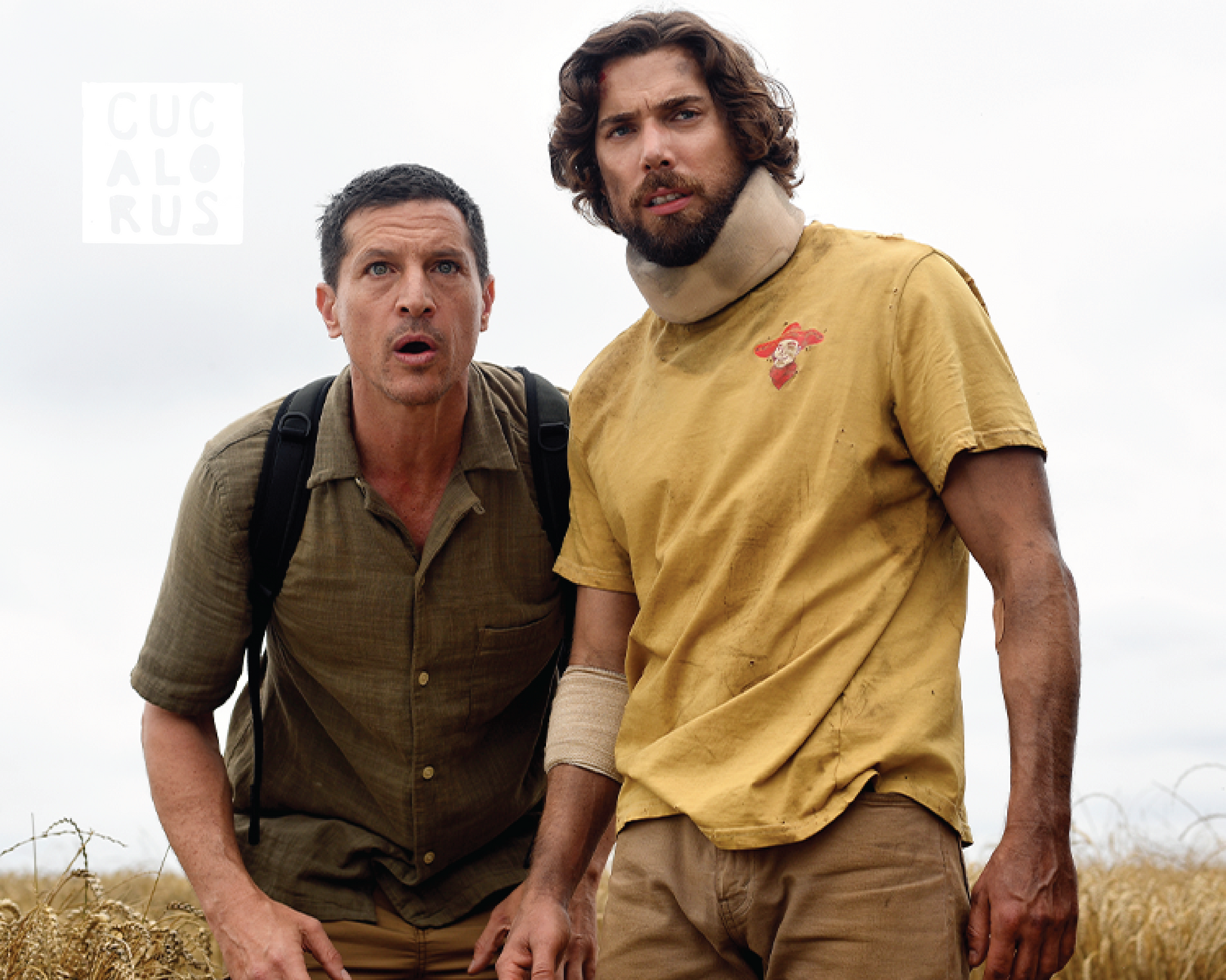 Two men crouch in a dry field under a bright sky, looking toward the camera.