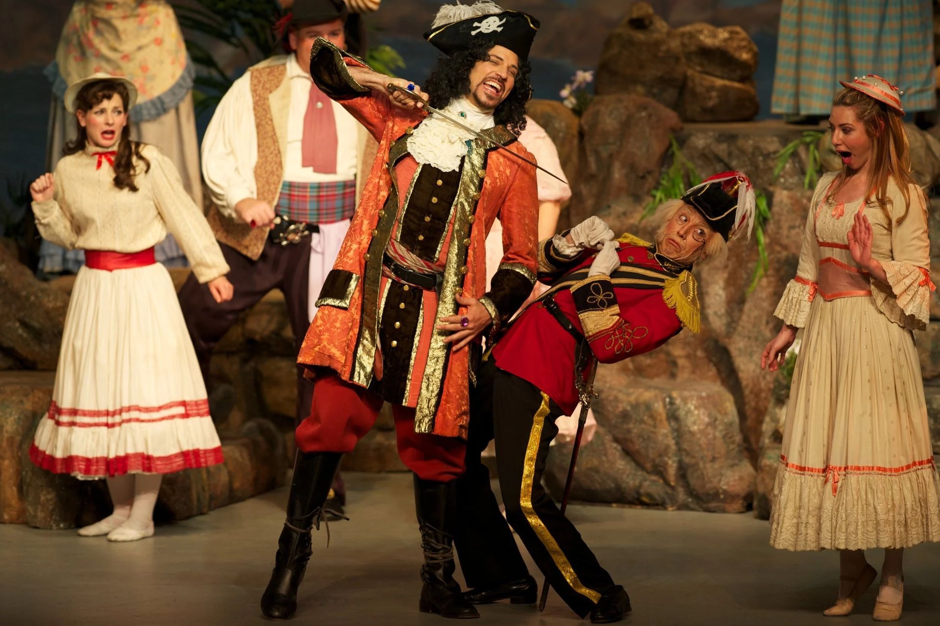 Costumed performers in a theatrical dance scene on stage, with one man in red and black at center.