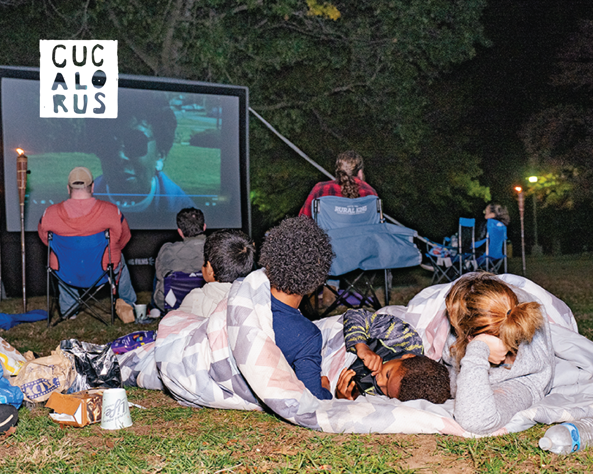 People on blankets watch an outdoor movie projected on a screen at night.