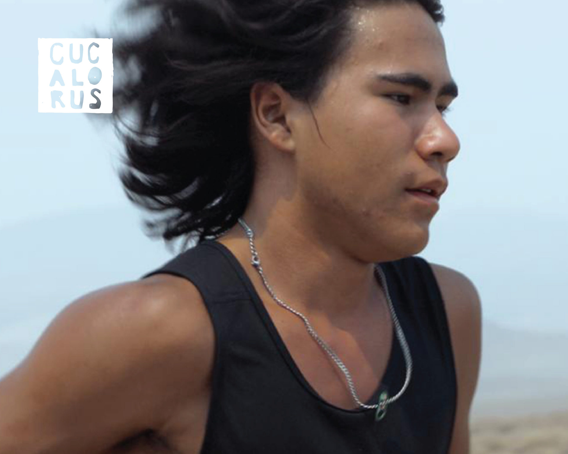 Person running on a beach with windblown hair, wearing a black tank top and silver necklace.