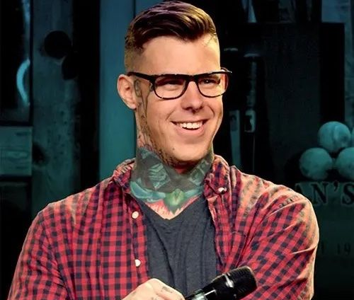 Smiling person in glasses and plaid shirt holding a microphone against a dark backdrop