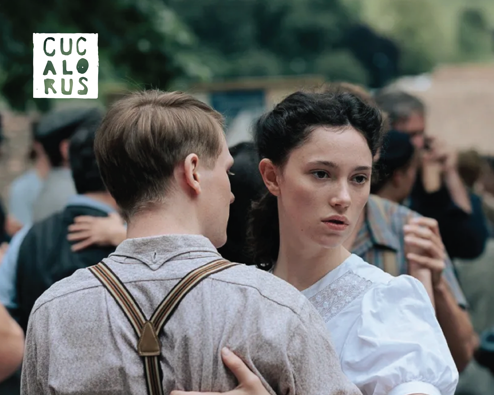 Two people in vintage-style clothing stand close together at an outdoor event, with a blurred crowd behind them.