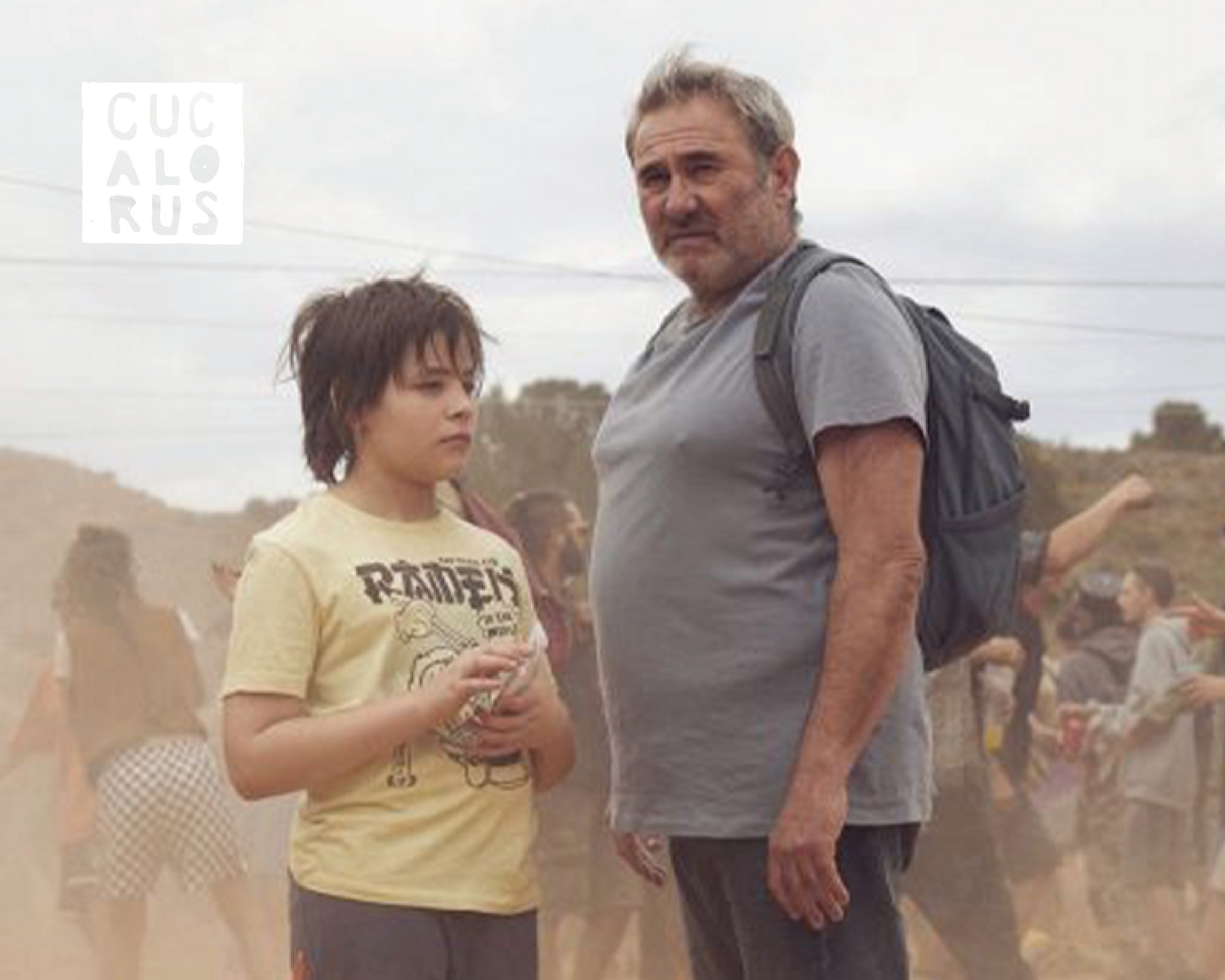 Two people stand in a dusty outdoor crowd, one in a yellow shirt, both facing the camera.
