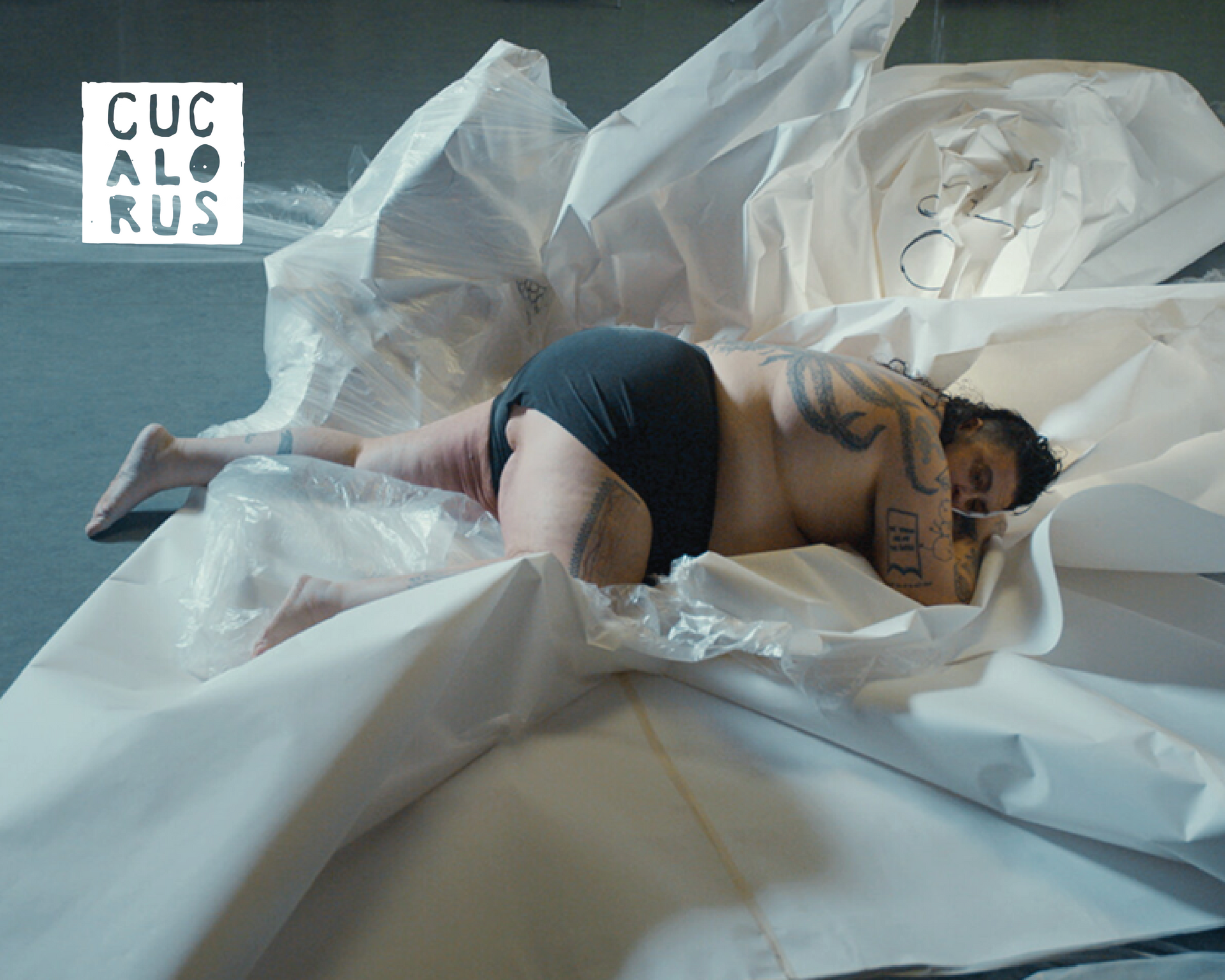 Person curled on white sheets in a soft-lit studio scene, with a small CUCALORUS logo in the corner.